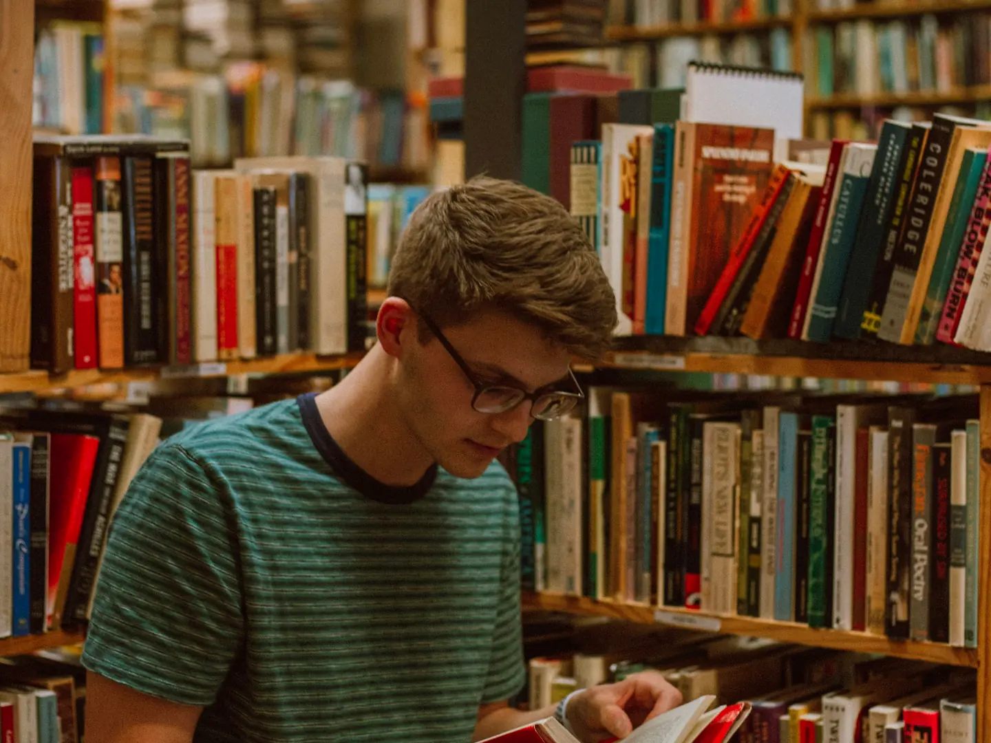 A young person wearing glasses reading a book beside library shelves