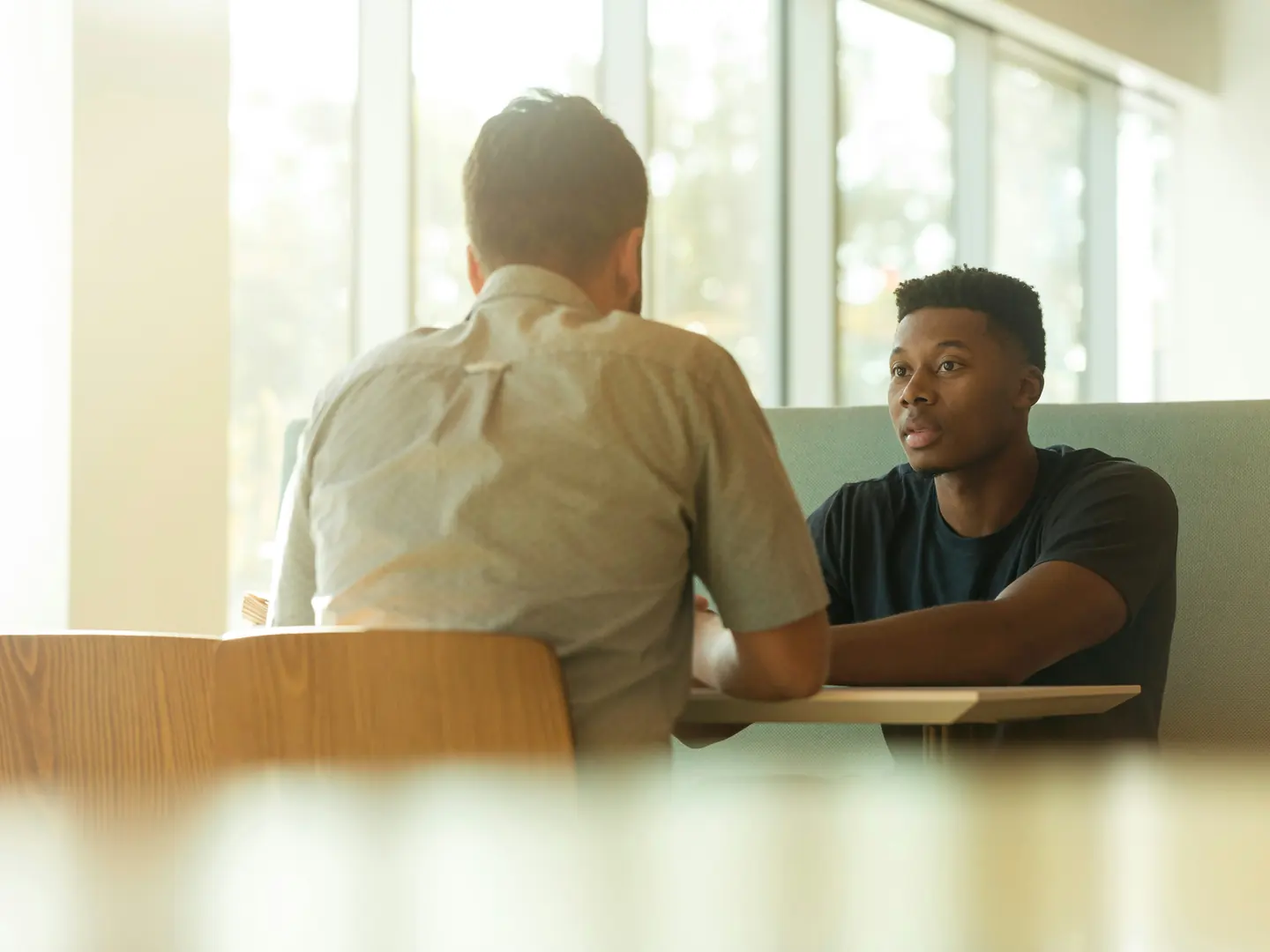 Two men seated across a table in conversation near a sunlit window